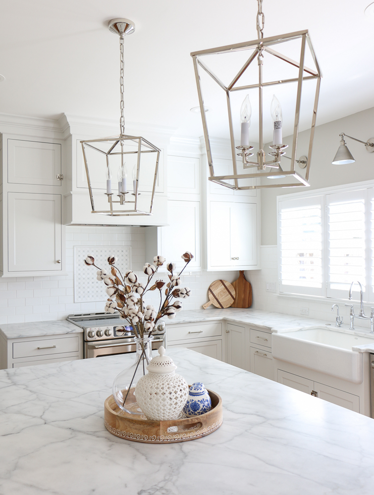 All white kitchen with polished nickel lanterns over kitchen island. Sink by window wall with wall sconces above