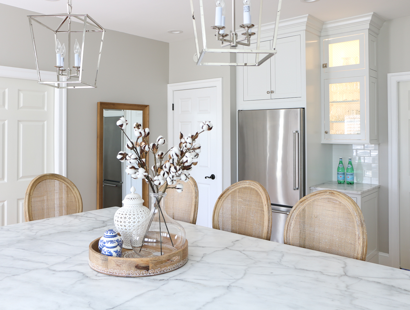 Sherwin Williams Agreeable Gray walls in kitchen with white cabinets, carrara marble kitchen island, cane back counter stools. Round tray centerpiece with ginger jars and glass vase