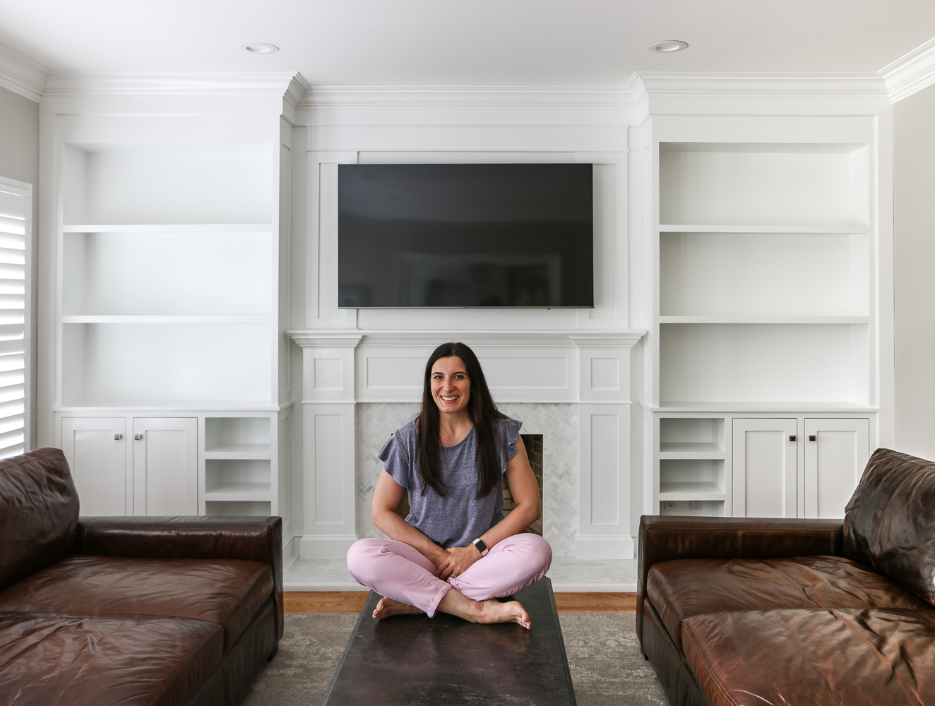 Stefana Silber sitting on coffee table in front of painted fireplace builtin project, open shelves without decor, leather sofas on each side of room