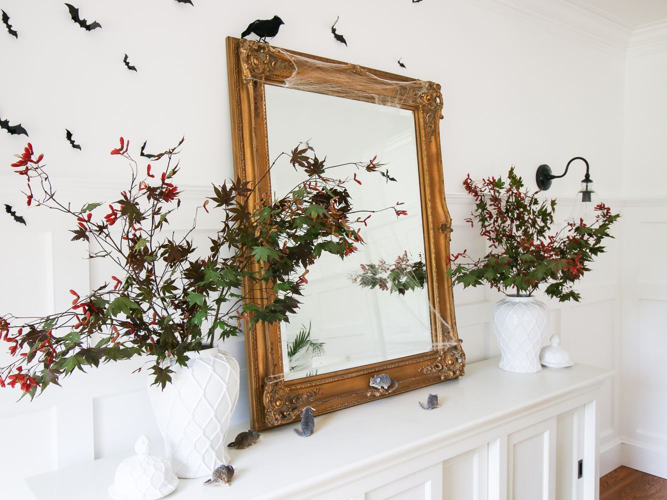 white dining room with white sideboard styled with vintage gold mirror, white ginger jars with Japanese maple branches, black bats on wall, blackbird and mice on console table