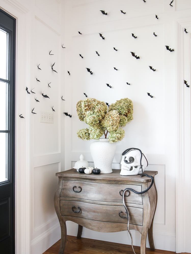 entryway console table with skull, snakes, and black bats on wall, white vase styled with dried hydrangeas
