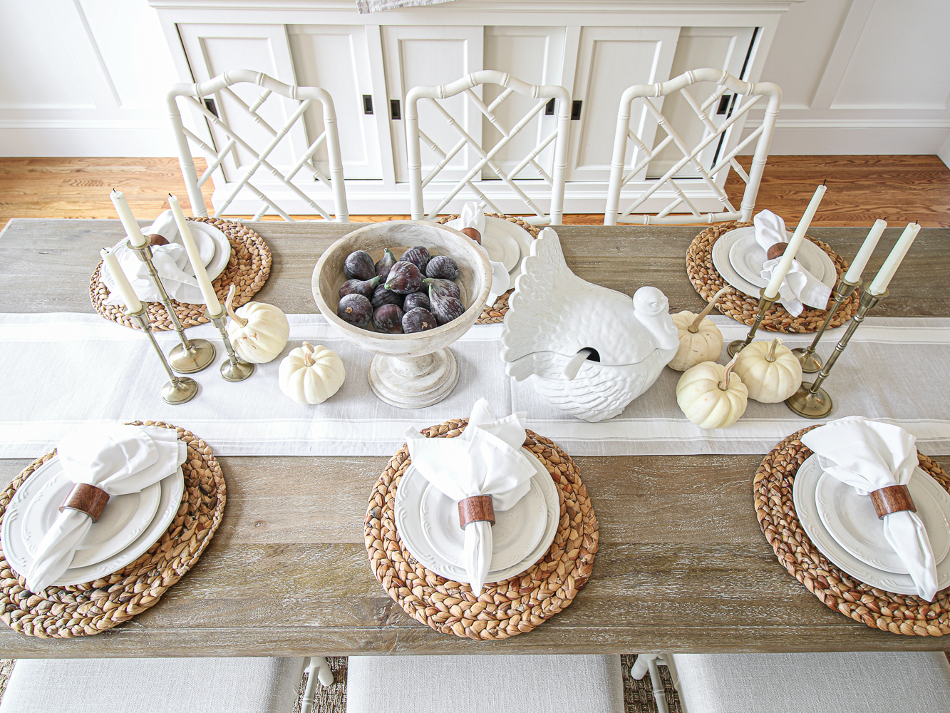 Thanksgiving table with white and neutral decor, turkey tureen, rectangle table, white dinnerware and napkins on top of woven chargers, white pumpkins, chippendale chairs, candles