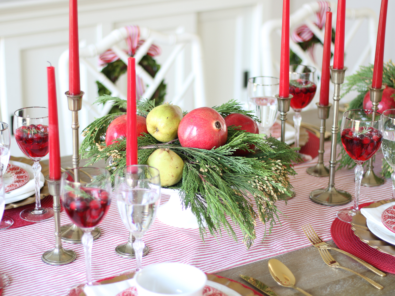 Christmas tablescape, red tapered candles, centerpieces with greenery, pomegranates and pears, crystal glassware with holiday cocktails, striped table runner, Chippendale chairs with boxwood wreaths