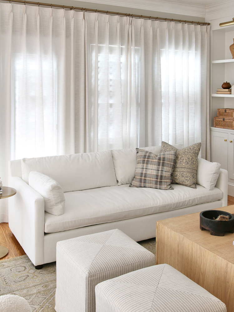 White linen pinch pleat drapes, white velvet West Elm sofa, square ottomans, hardwood floors, builtin shelves styled with decor