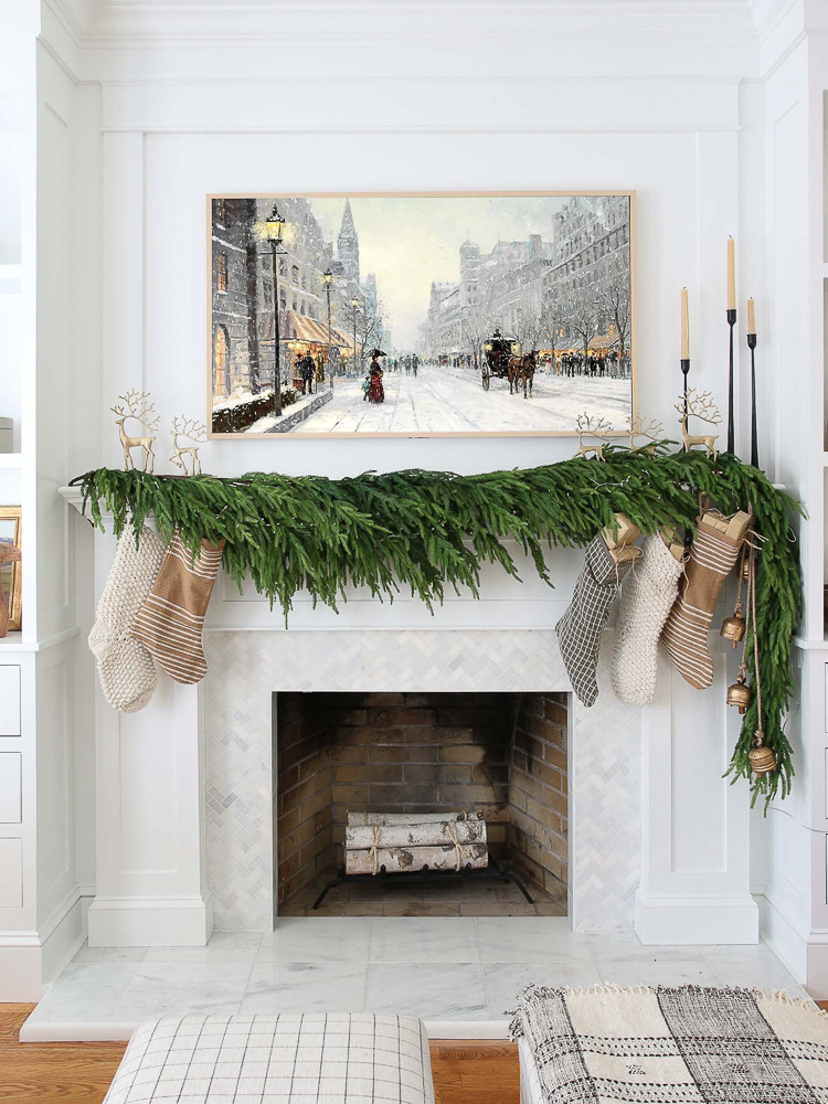 fireplace mantel with 5 faux Norfolk pine garlands, frame TV with vintage art, marble fireplace surround, assorted stockings, bells, gold reindeer, iron cabdle holders