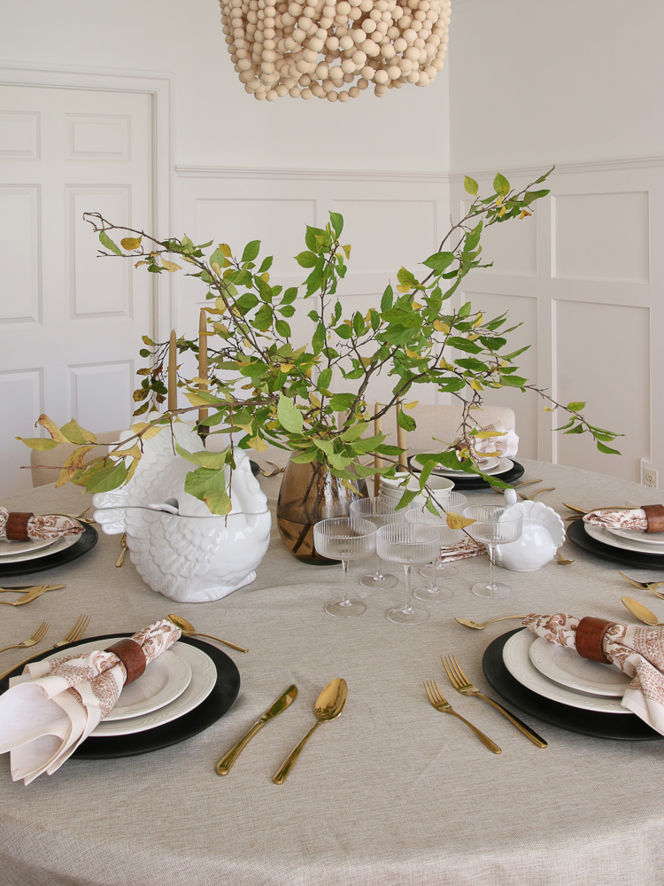 Round table setting for Thanksgiving, turkey soup tureen, white dishes, gold utensils, linen tablecloth, centerpiece greenery