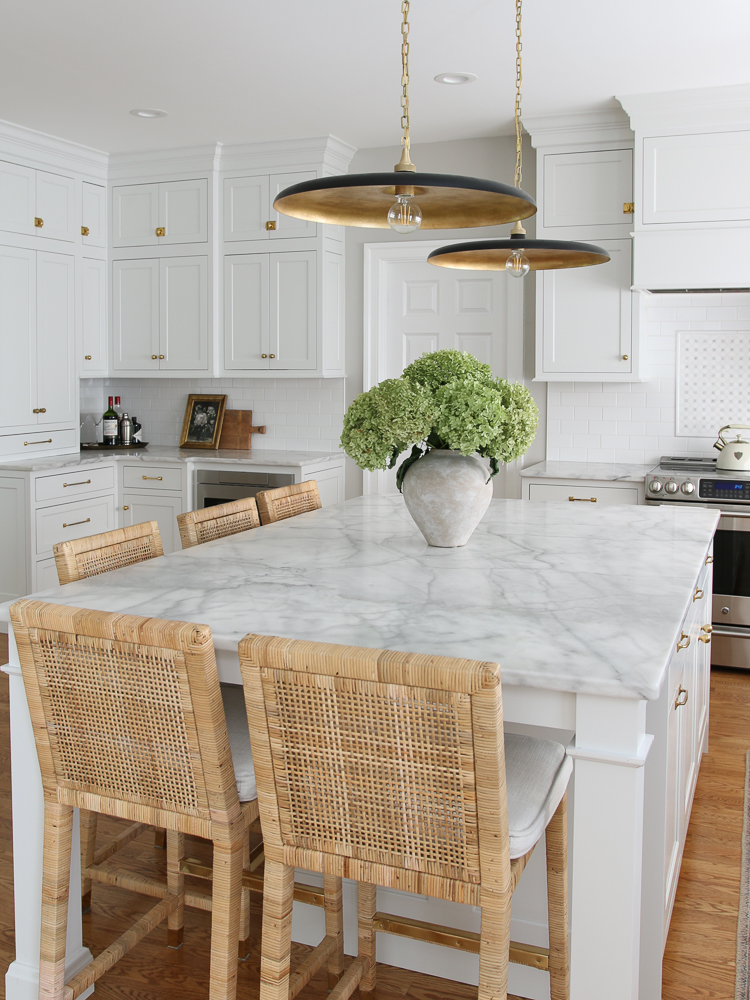honed marble countertops in all white inset cabinets kitchen, white subway tile backsplash, black and gold pendant lights, woven counterstools