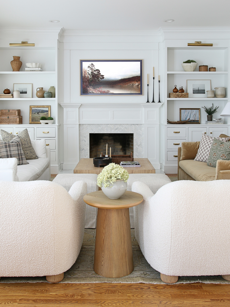 view of white living room with pair of white boucle chairs, two velvet sofas, fireplace all with builtins around tv