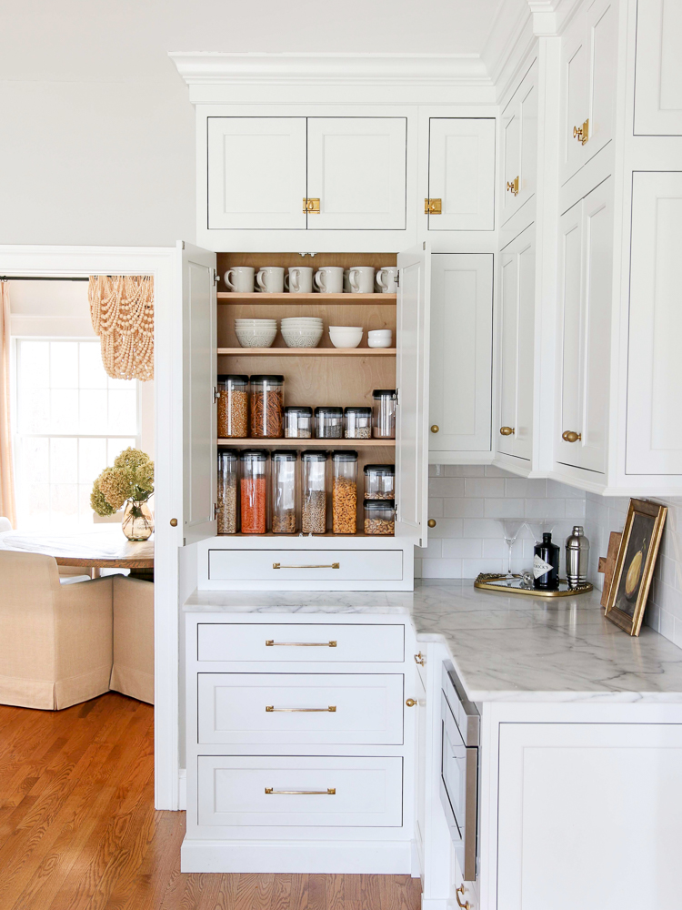 white kitchen with pantry cabinet storage, organized with clear food containers for snacks and cereal, bowls and mugs on upper shelves, dining room seen in background, honed marble countertops, white subway tile backsplash,  unlacquered brass hardware