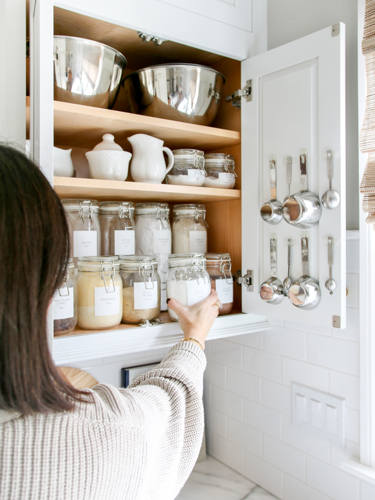 woman reaching into organized pantry cabinet for dry baking goods stored in air-tight glass jars, measuring spoons hung on inside of cabinet door to maximize space