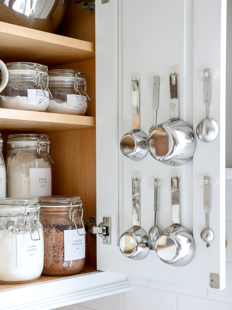 pantry cabinet open with organized baking goods and supplies, metal measuring spoons hanging on inside of door with command hooks