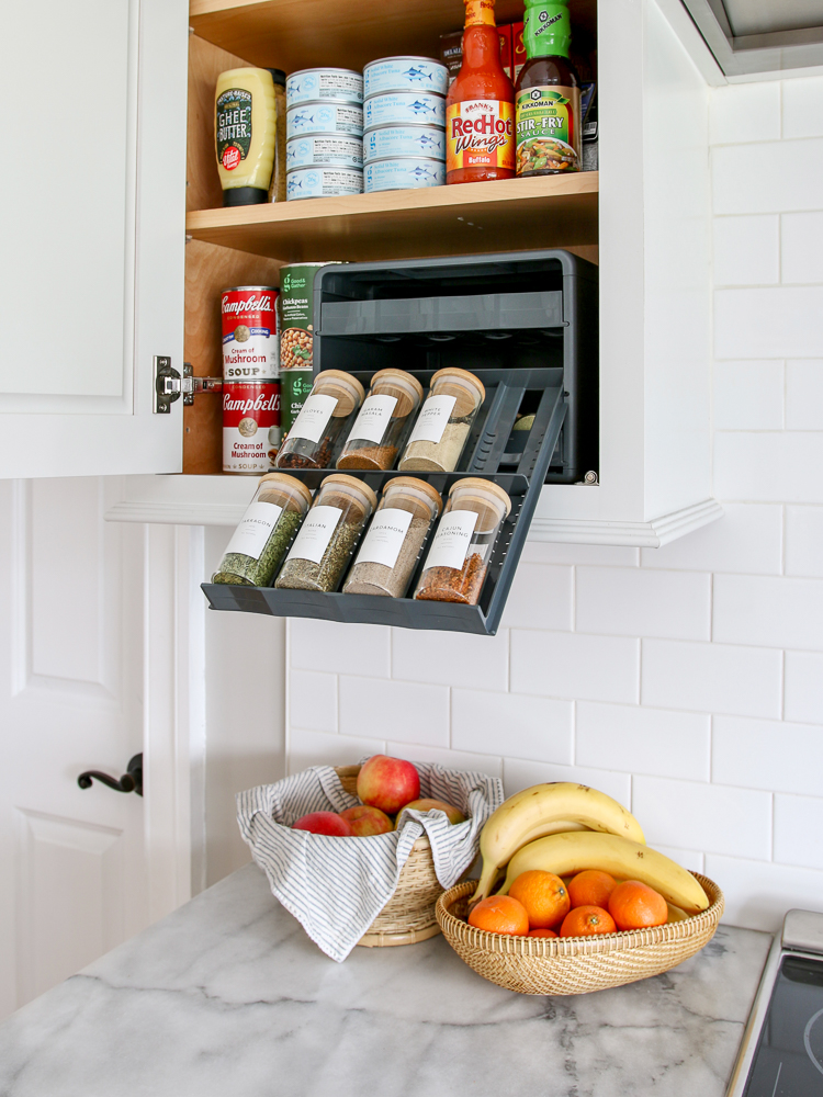white kitchen pantry cabinet open with pull out spice organizer showing spices, canned goods and sauces in cabinet, white Carrara marble countertops with fruit baskets, white subway tile backsplash