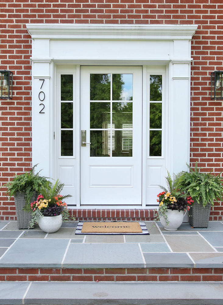 red brick home with bluestone front porch and white door, planters with flowers on either side