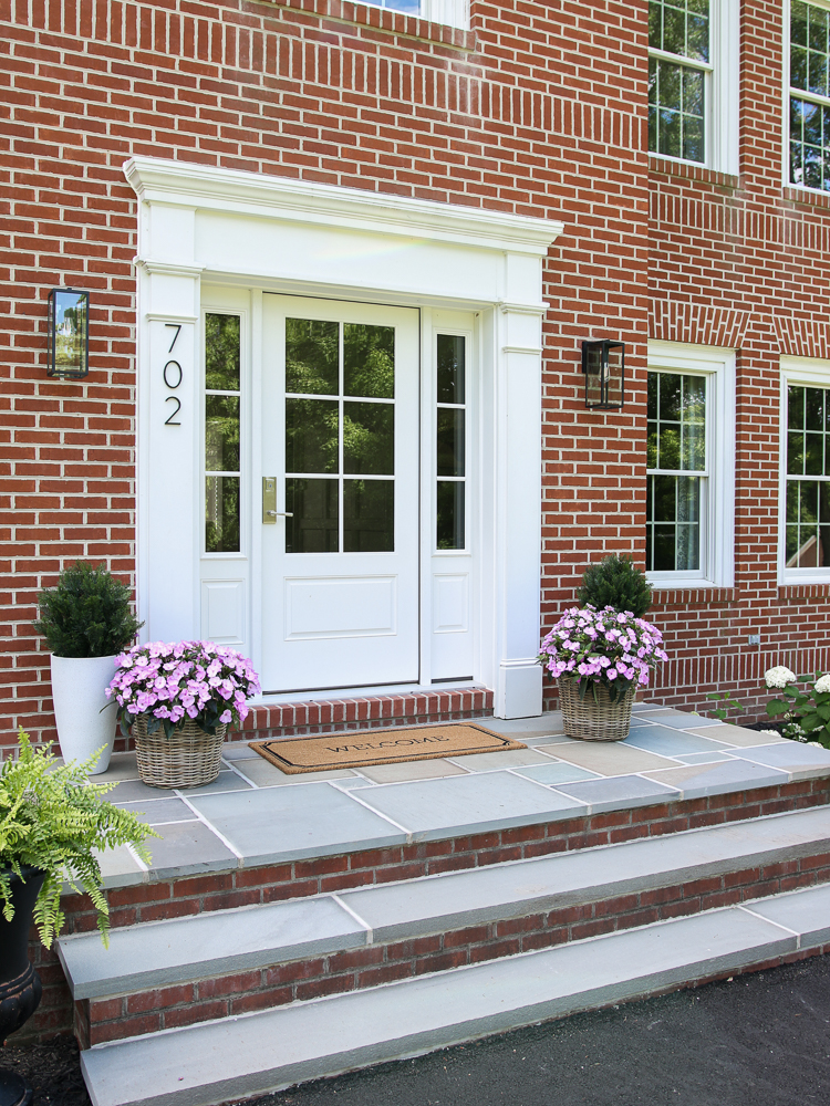 after view of brick home with bluestone porch, flowers, and welcome mat