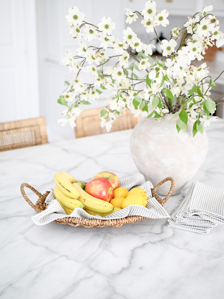 Carrara Marble countertop on island, basket with bananas, apples, and lemons, vase with white flowers