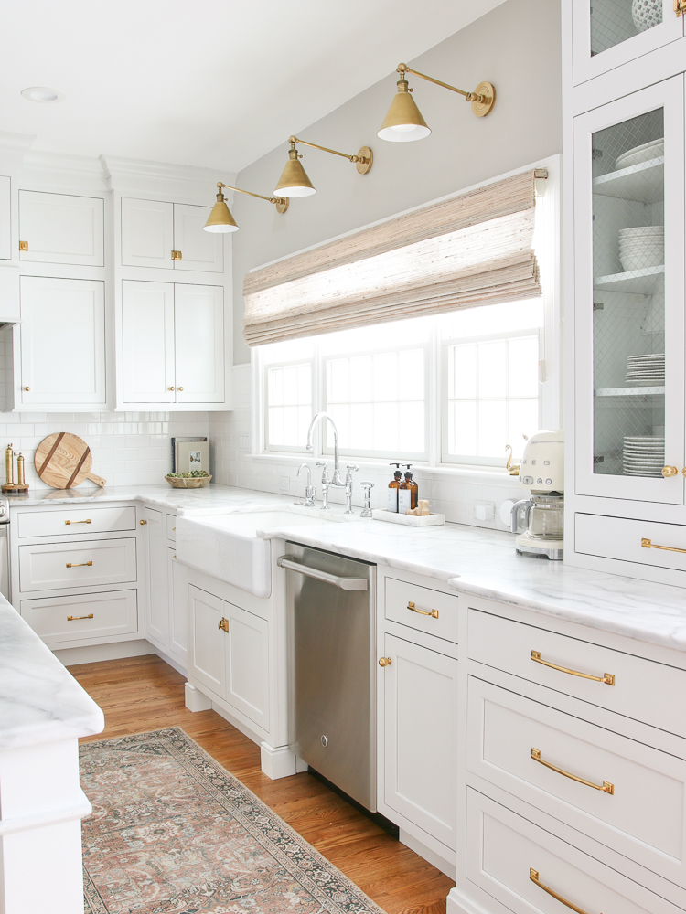 window above classic white kitchen sink with woven shade and three brass sconces above window