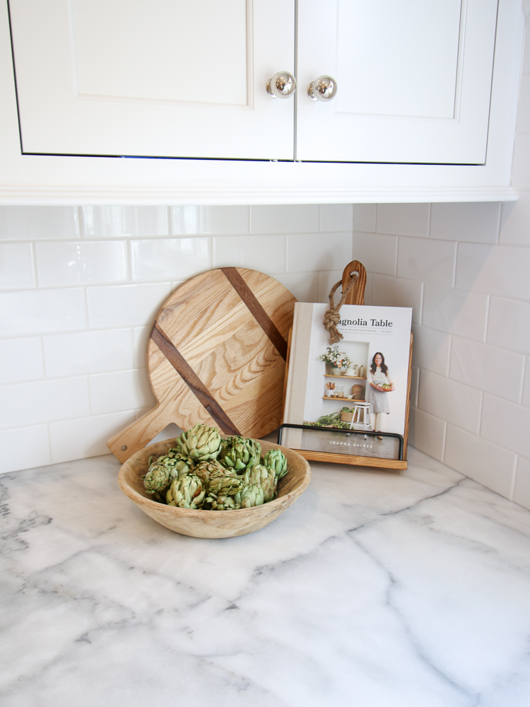 corner of kitchen countertop with cookbook on display, round wooden cutting board, wooden bowl with artichokes, white kitchen cabinets