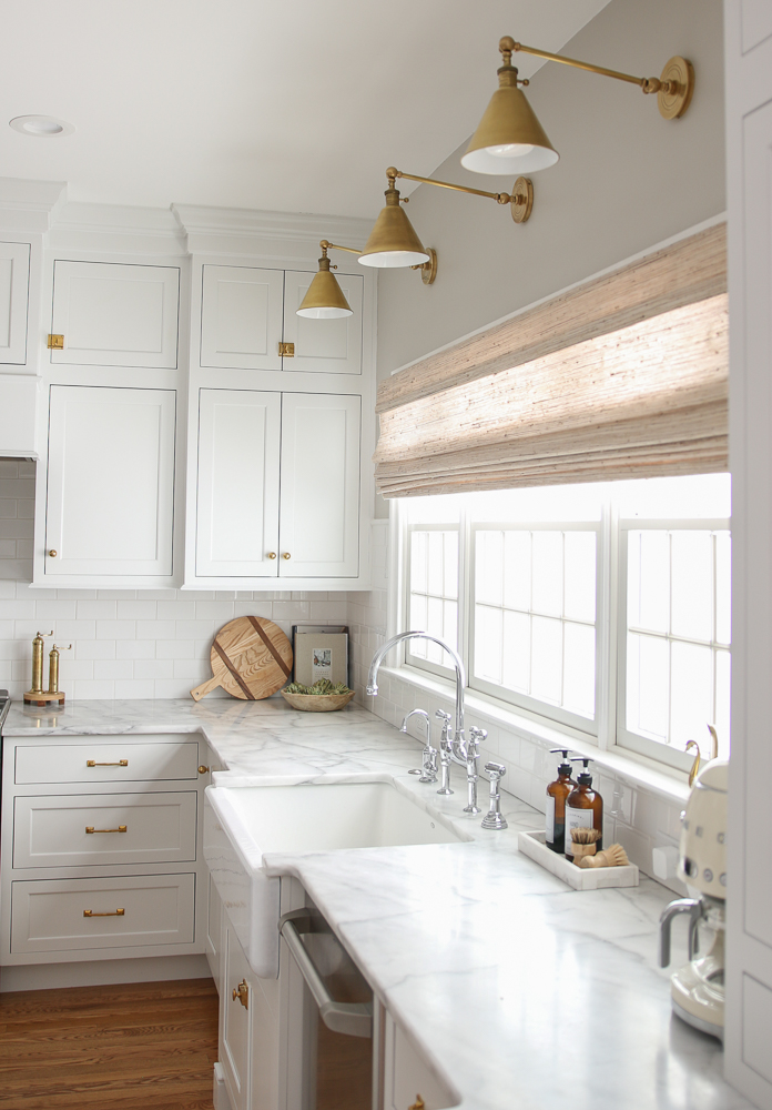 white kitchen with woven shade over window, brass sconces and cabinet hardware