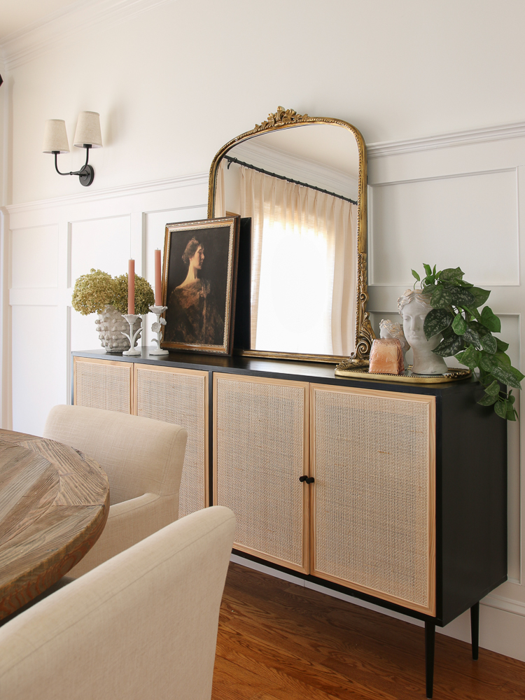 Gleaming Primrose mirror styled over a black sideboard with woven doors, artwork, candles, plants, wall molding, sconces