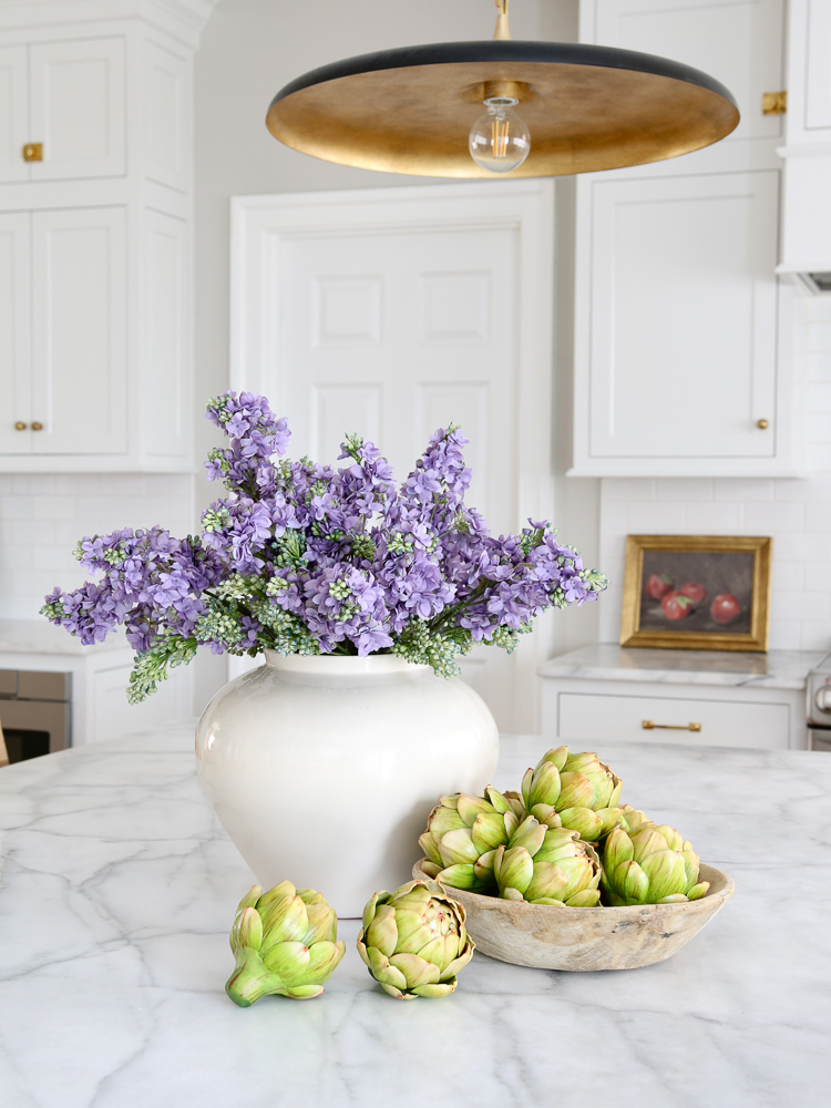 faux lilacs in a vase on a kitchen counter next to a bowl of faux artichokes