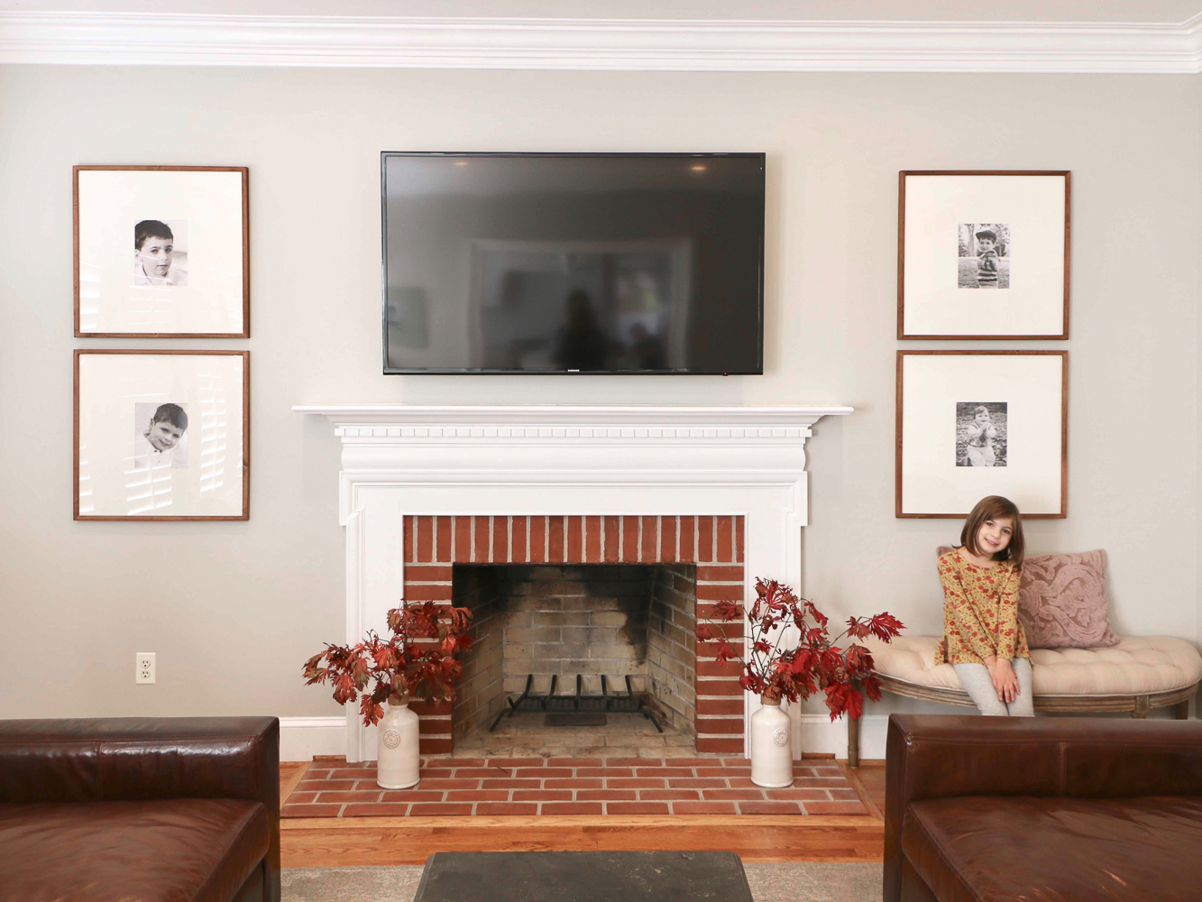 Brick fireplace with oversized framed family photos and tv above fireplace, tufted demilune bench on right side. Wall paint - Sherwin Williams Agreeable Gray