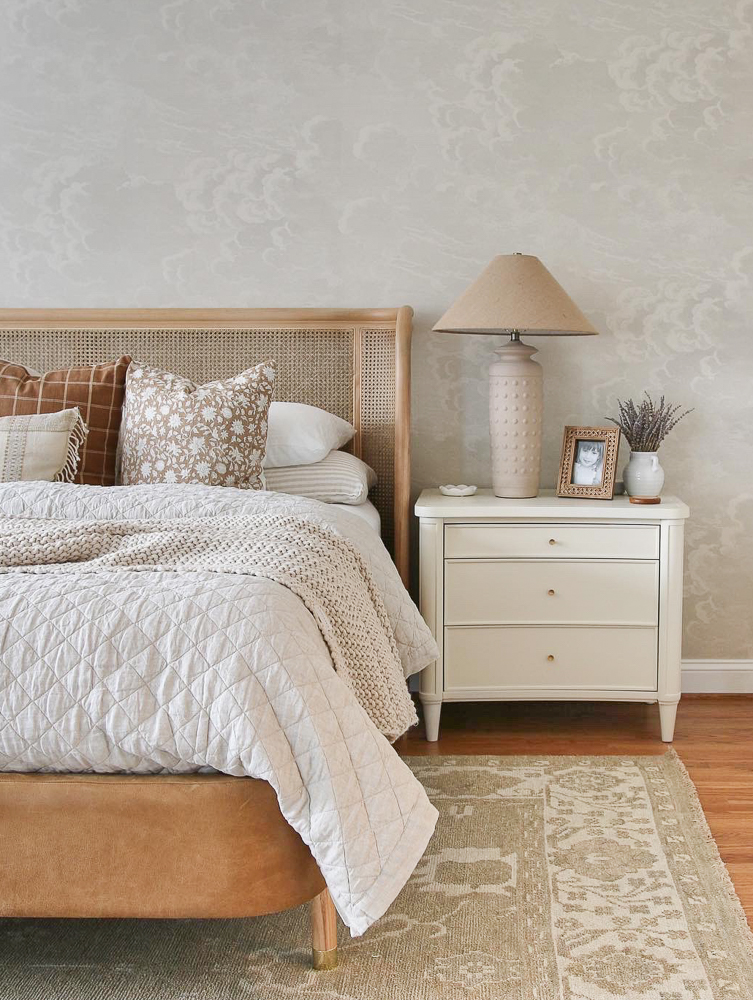 primary bedroom with neutral wallpaper, bed with woven headboard and light colored neutral bedding, white nightstand with neutral colored lamp and decor