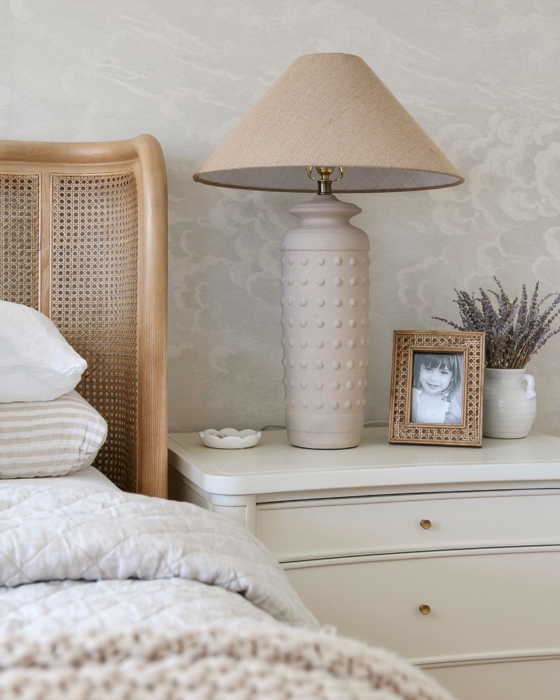 close up showing side of cane bed and white nightstand decorated with lamp picture frame and floral arrangement against neutral wallpaper in the background
