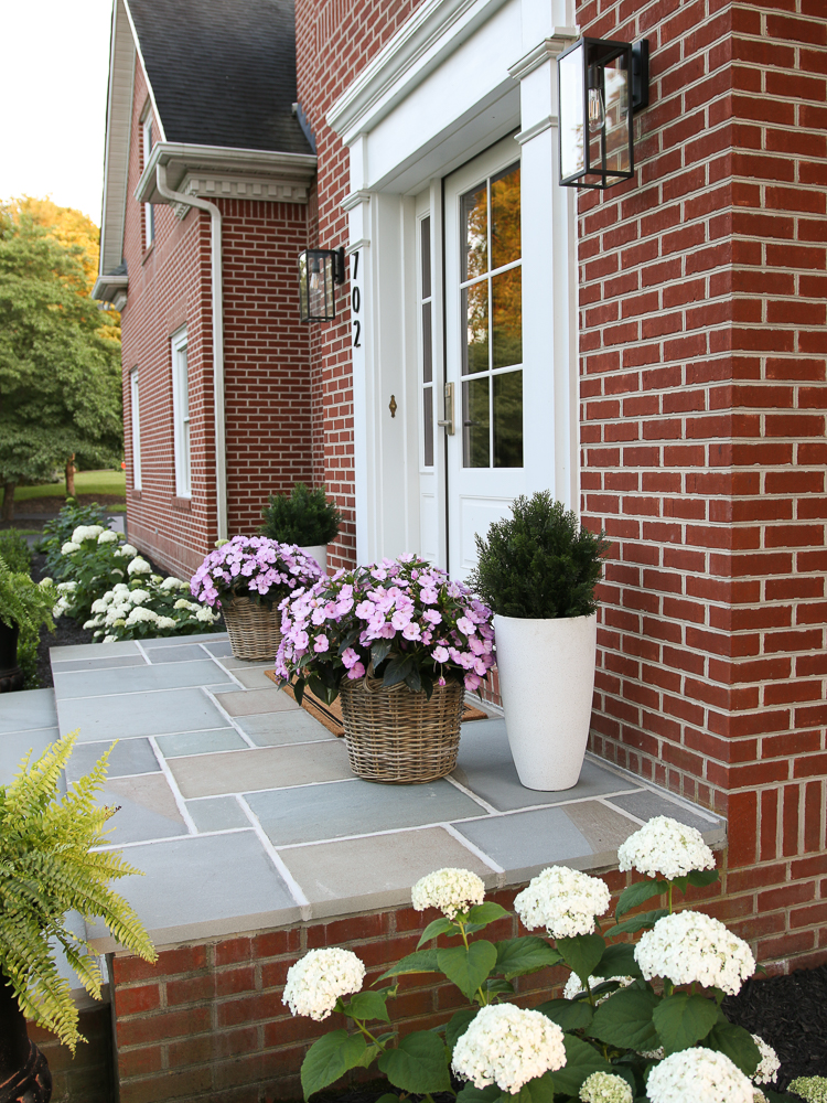 side view of bluestone front porch with no roof, red brick home with white front door with basket and planters on either side, pink flowers and greenery, hydrangeas