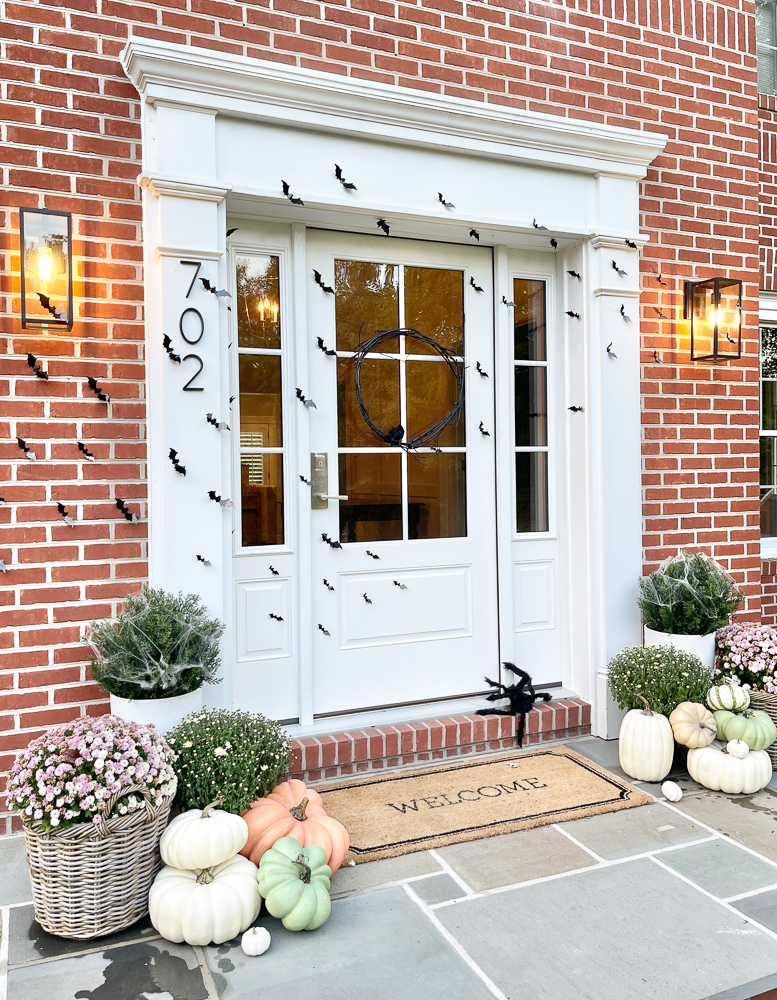 fall and Halloween decorations in front of red brick home, small bats on front door, welcome mat with planters with mums, and assortment of light colored faux pumpkins