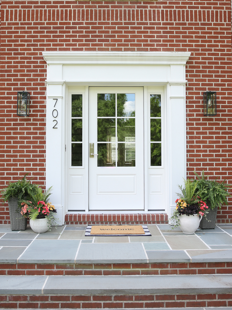 brick home with white front door with windows, open porch with bluestone surface, welcome mat, planters on either side of door with fern and a mix of flowers