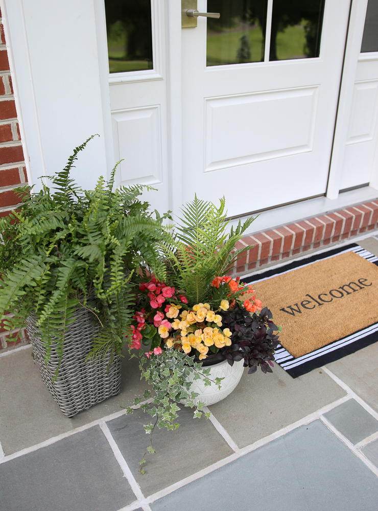 close up of uncovered front porch with bluestone surface, welcome mat, planter with mix of red and yellow flowers and greenery, tall gray woven planter with fern