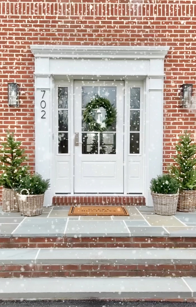 snowy winter porch decor, artificial shrubs and trees on either side of front door in woven planters, red brick home with white front door and wreath