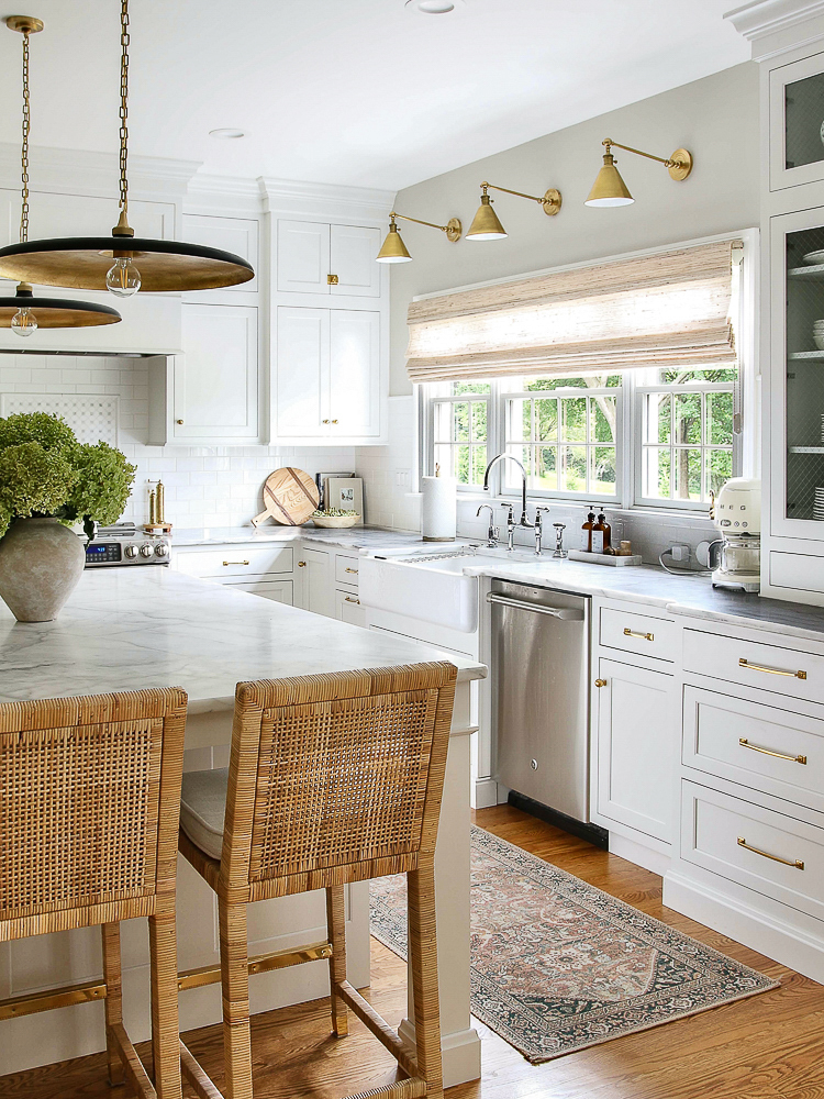 classic white kitchen with unlacquered brass hardware, Carrara Marble countertops, brass sconces over the sink window with woven shade, black and brass pendants over island