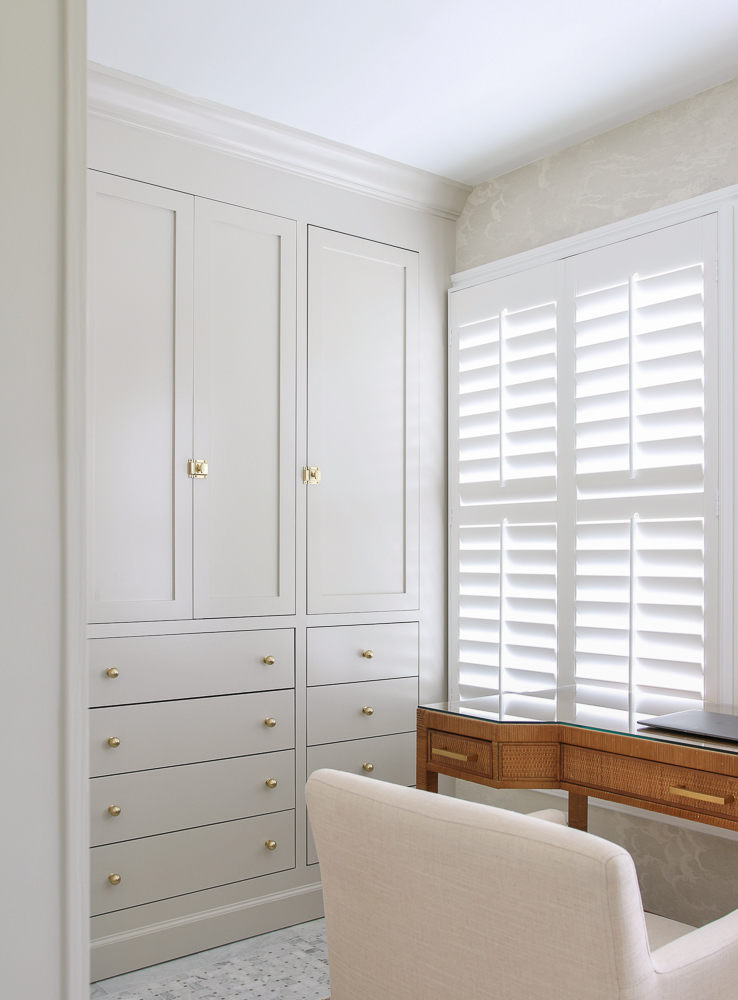 Corner view of primary closet with marble floors and IKEA PAX hack to look like custom cabinetry painted accessible beige, brass cabinet hardware, window with white interior shutters, brown woven desk with  linen chair
