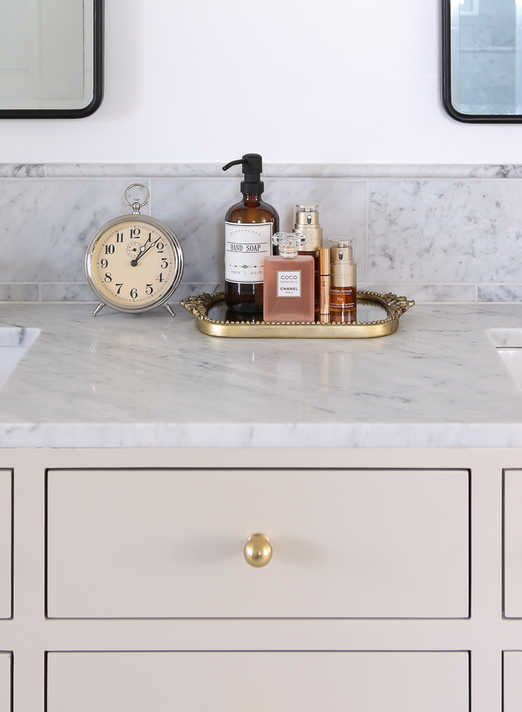 close up of vanity decor, gleaming primrose tray with hand soap, perfume and beauty products, white wall, vanity with marble countertop and brass hardware, black framed mirrors