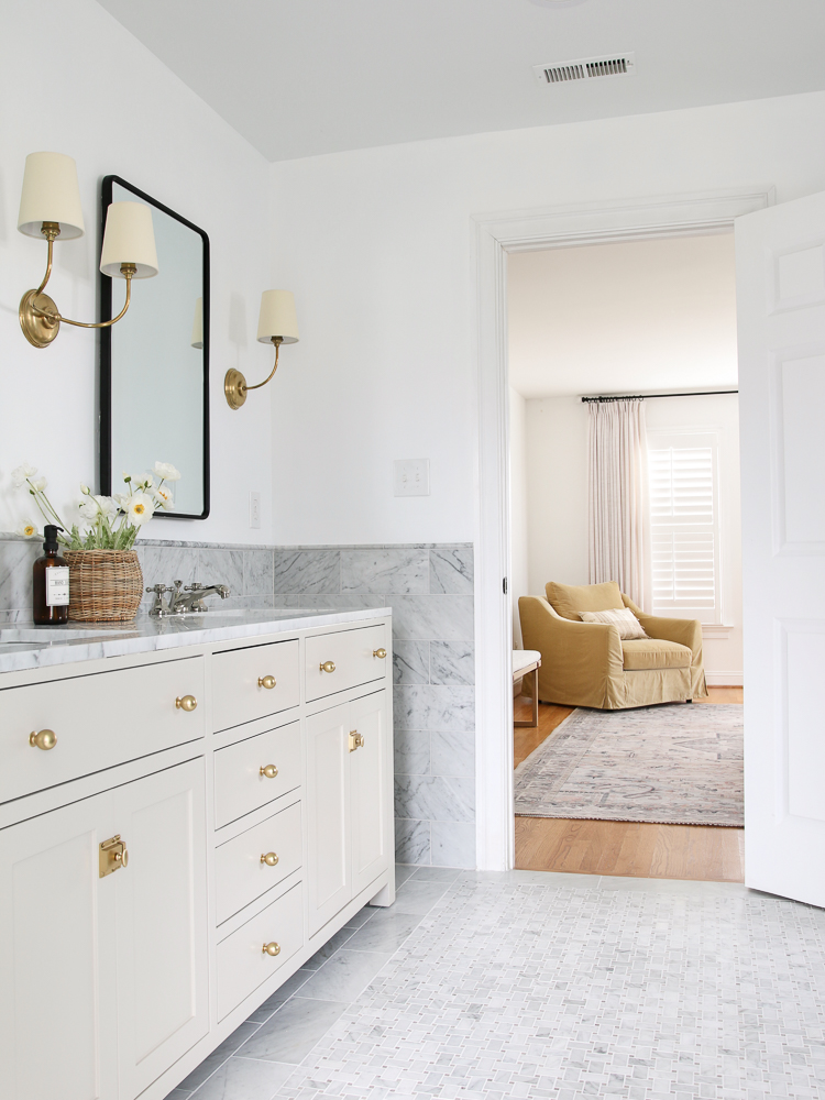 View from bathroom into primary bedroom, marble tile on floors, backsplash wall, and countertop. Brass cabinet hardware on Accessible  Beige painted vanity. Bedroom has hardwood floors and a camel colored chair in the cornier by a window. 