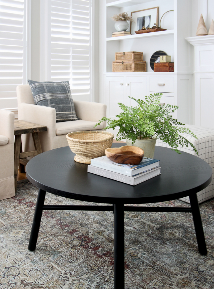 black round coffee table with stack of 2 books, woven bowl, and small plant, built-in bookshelf in background with decor