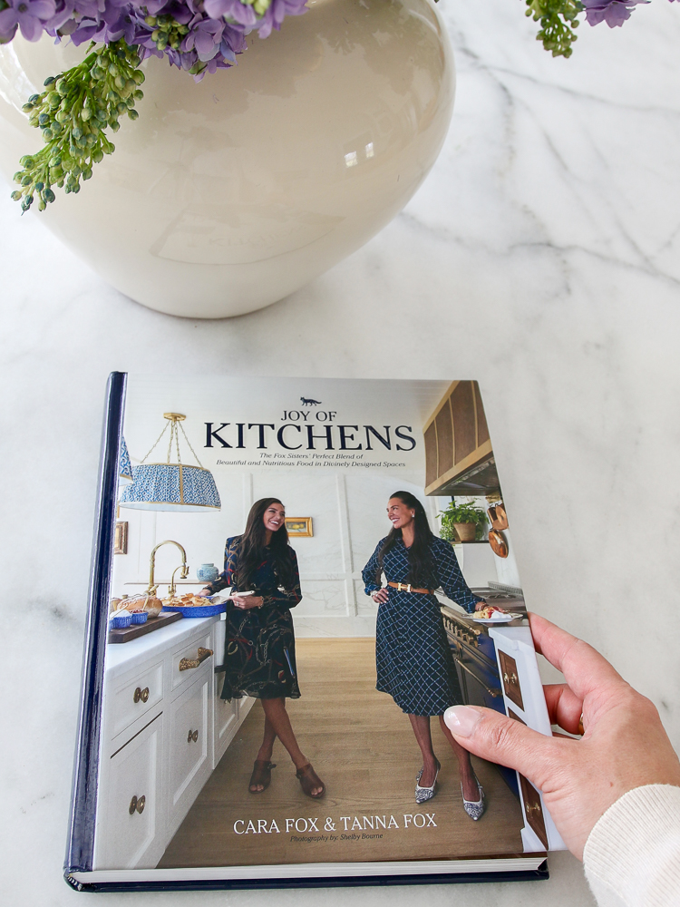 Stefana Silber holding coffee table book about kitchens on marble countertop next to vase with faux lilacs
