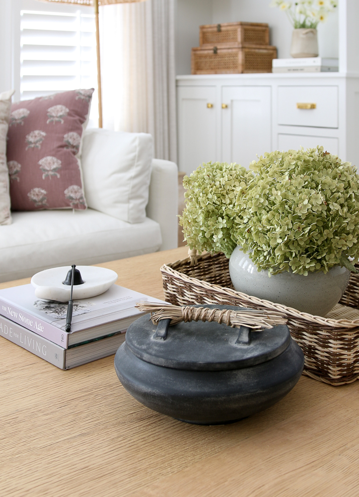 close-up of home decor gifts styled on the coffee table including a black Anthropologie trinket dish, home decor books, candle snuffer, and a vase with dried hydrangeas, white velvet sofa in background with built-ins shelves 