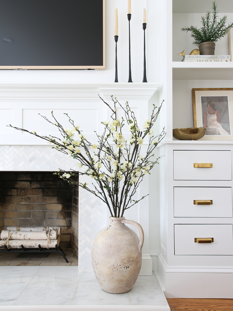 large artisan vase on the floor next to the marble tile fireplace with faux branches that have pale yellow flowers, white built-ins with brass cabinet hardware