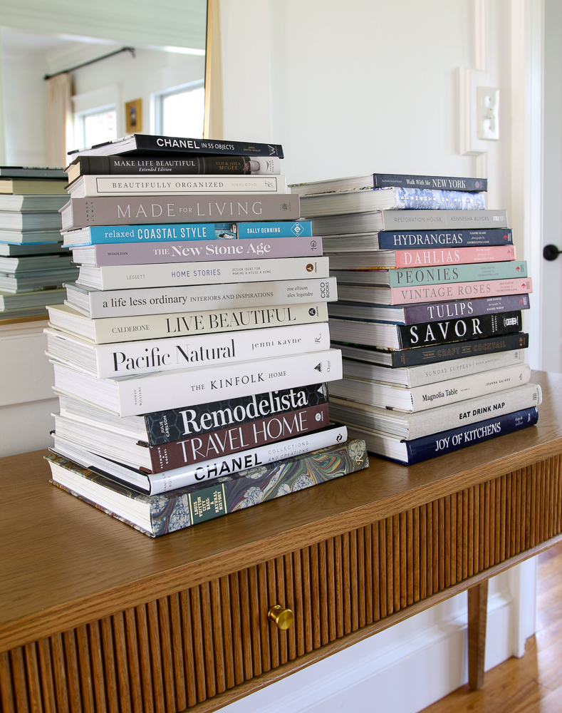 two large stacks of coffee table books on a wood console table in front of a brass frame mirror, examples of home decor gifts