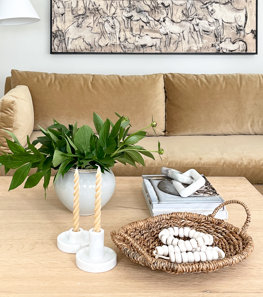 neutral living room with velvet sofa, wood coffee table with decor including books, marble geometric chain links, woven basket, wooden bead garland, marble candle holders with a pale yellow tapered candle, vase with greenery