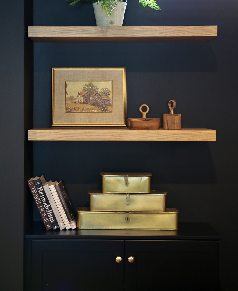 stackable brass boxes styled on a light wood shelf next to several vertical coffee table books, walls painted black
