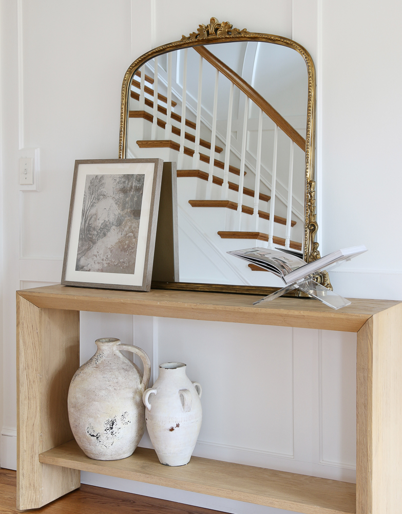 light wood entry console table in the hallway with white walls and wood floors, styled with gleaming primrose mirror, artisan vases, an open coffee table book on an acrylic book holder, and wall art