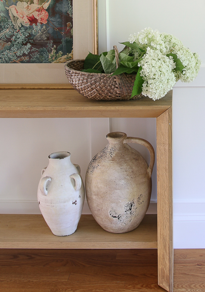entry table styled with artwork and a basket with fresh cut hydrangeas, two artisan vases on the bottom shelf