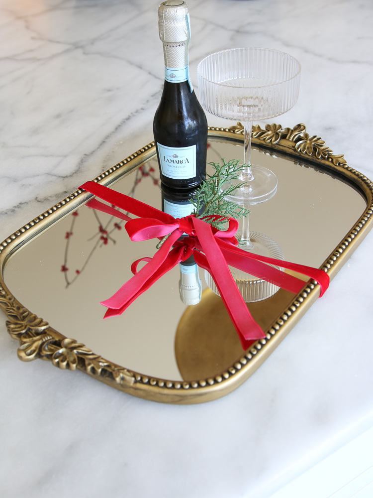 gleaming primrose mirror being shown on a marble countertop with red ribbon, a sprig of greenery, Prosecco  bottle, and fluted glass as home decor gifts