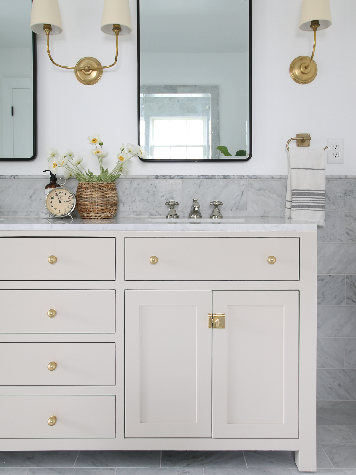 painted bathroom vanity cabinet in SW accessible beige with a basket of flowers on top. Black metal framed mirrors and brass sconces. Marble tile wainscoting on walls