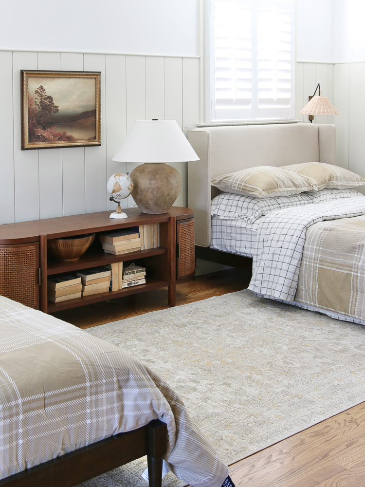 angled view of beds in boys room, headboard with with separate wood frame, vertical shiplap, console table and ceramic lamp, neutral beige. bedding and rug