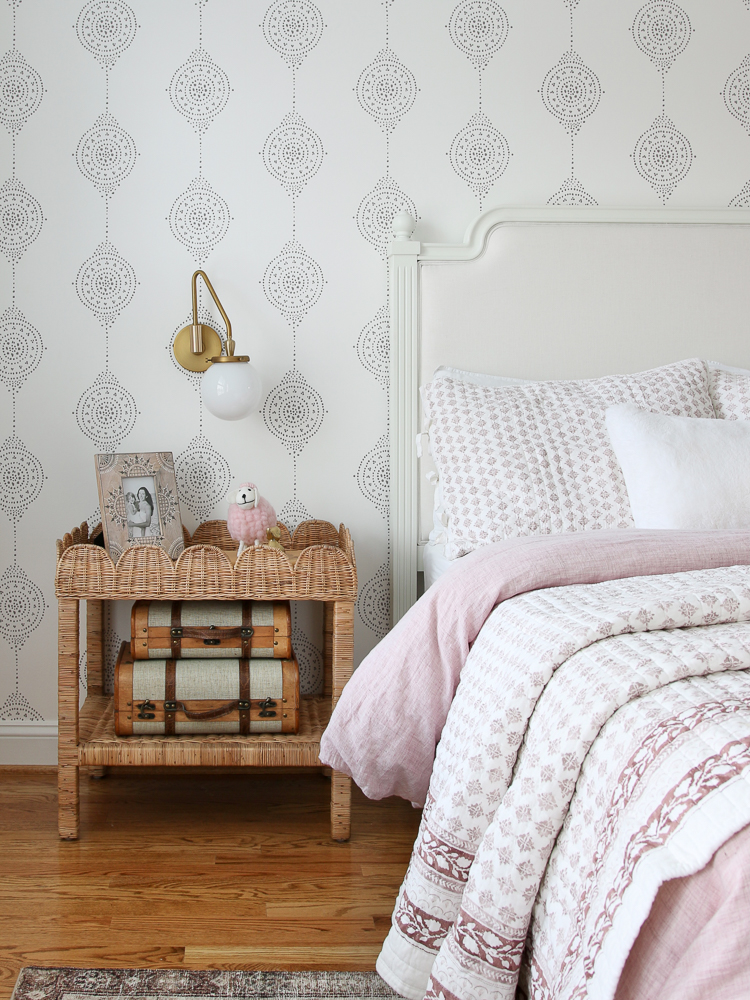 view of side of bed and woven nightstand, white wood and fabric from pottery barn, Serena and Lily wallpaper, pink and white bedding, hardwood floors and area rug