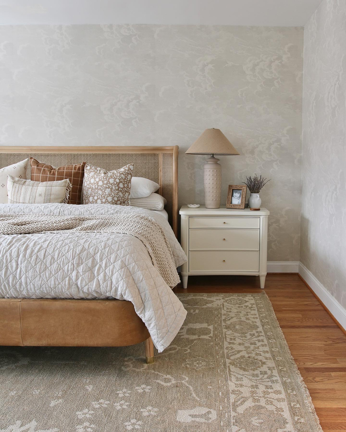 Bedroom with neutral cloud wallpaper and cane headboard. Linen quilt and plaid and floral throw pillows. White painted nightstand with tall hobnail ceramic lamp with burlap tapered shade. Vintage hand knotted rug